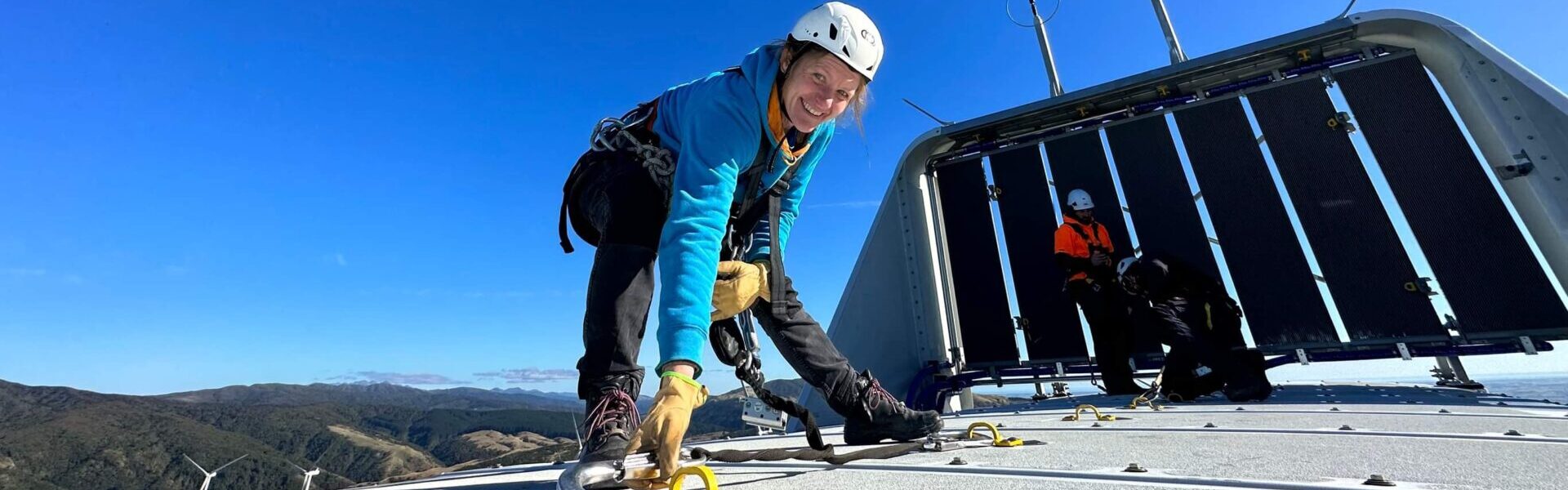 A woman smiles on top of a wind turbine with helmet and harness and a blue sky in the background