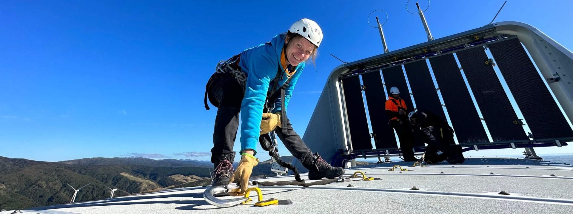 A woman smiles on top of a wind turbine with helmet and harness and a blue sky in the background