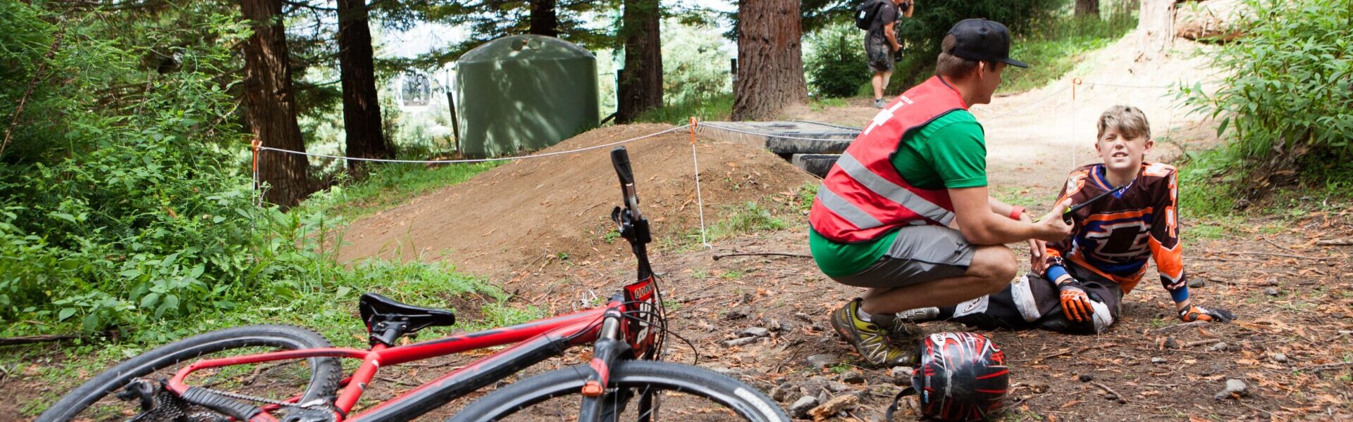 A man with medical attire crouches next a boy sitting on the forest floor with a bike on its side in the foreground