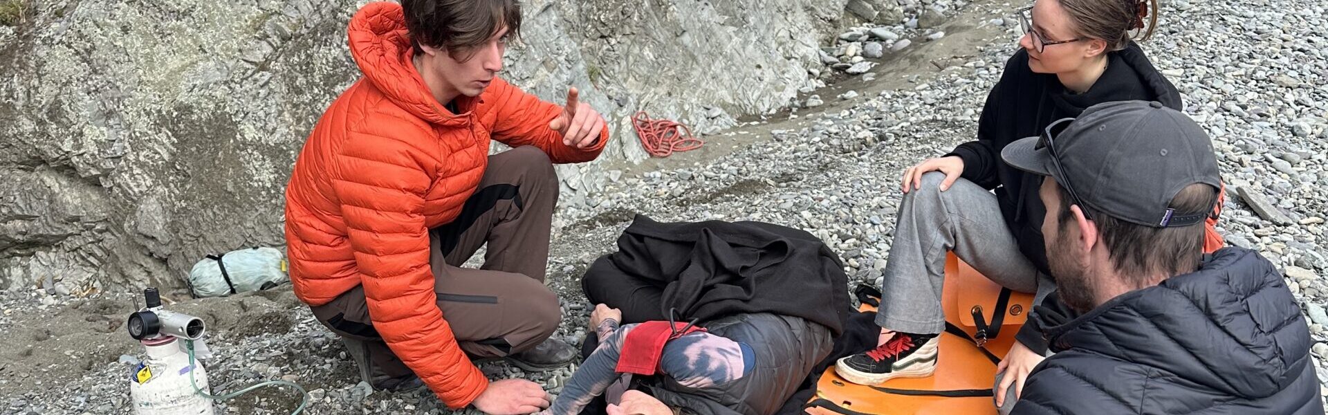 Three people sit on a river bank with a patient lying between them