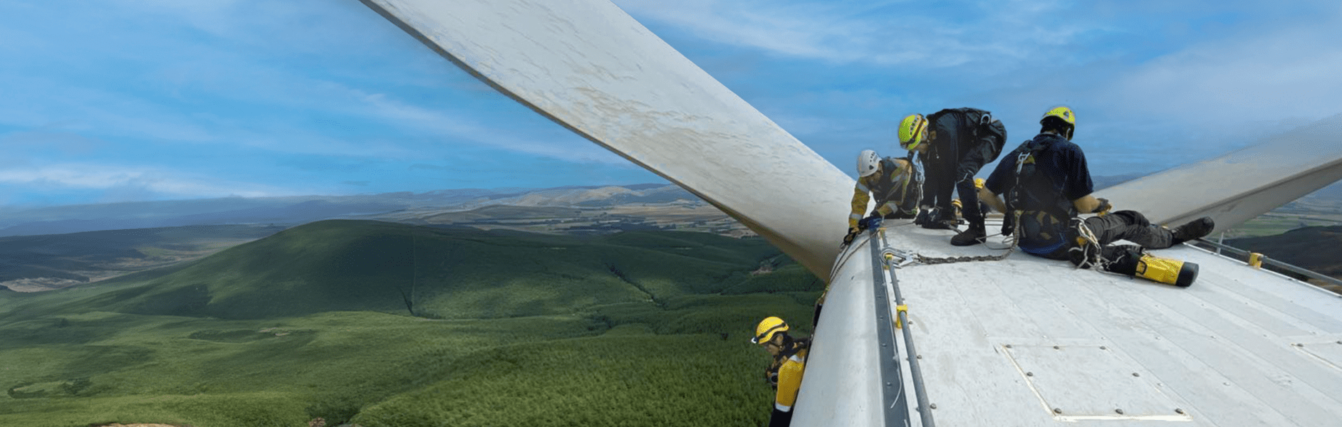 Three people in safety harness sit on top of a wind turbine